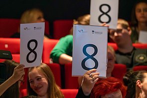 Publikum im Spreekino Spremberg bei der Abstimmung nach einem Slam. Die Gäste halten DIN A4 große Abstimmungskarten nach oben. Alle eigen die Bewertungszahl 8.